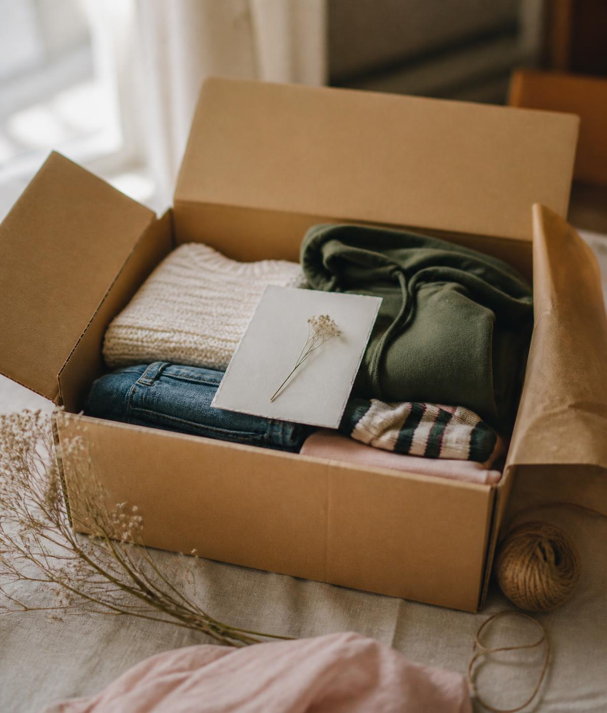 An open kraft box packed with folded clothes — cream knit sweater, olive hoodie, denim and striped socks — with a handwritten note on top